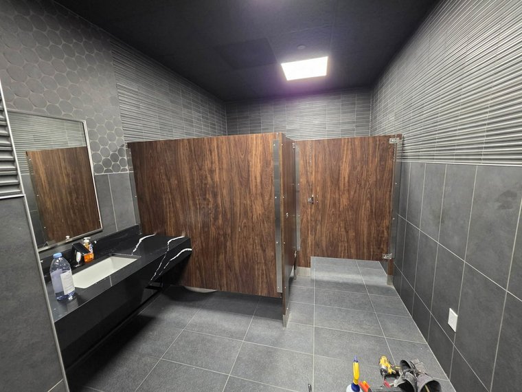 Finished commercial restroom with wood-grain partitions, gray tile walls and a quartz vanity